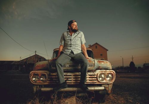 A bearded man in a patterned shirt sits on the hood of a vintage car in a dusty town at sunset, looking off into the distance.