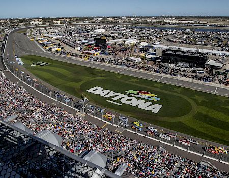 Aerial view of a NASCAR race at Daytona with a large crowd, green infield grass, a prominent Daytona logo on the infield, and the track lined with stands.