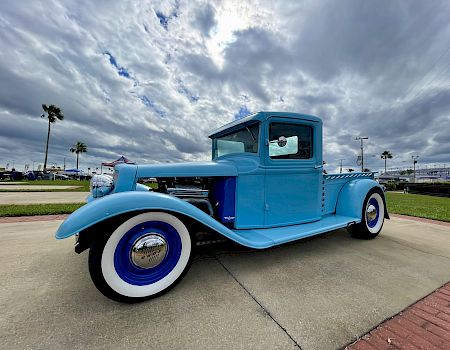 A vintage blue hot rod with whitewall tires parked on a sunny day, classic car lines, open front fender, and a dramatic cloudy sky behind it.