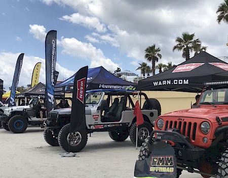 Off-road vehicles lined up on a sandy beach, with tents and banners, showcasing rugged Jeeps and side-by-sides under a sunny sky.