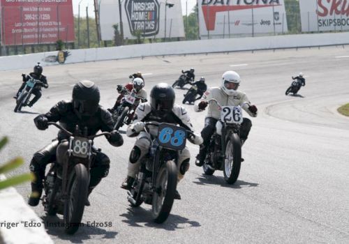 A pack of vintage motorcycles races around a track, leaning into a bend, riders wearing helmets; speedway-style competition on a sunny day.