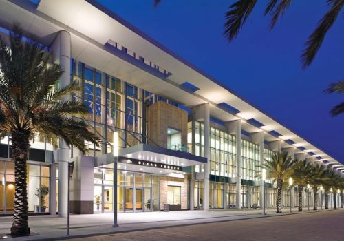 A modern glass-front building at dusk, with palm trees lining a lit walkway and a wide entrance canopy.