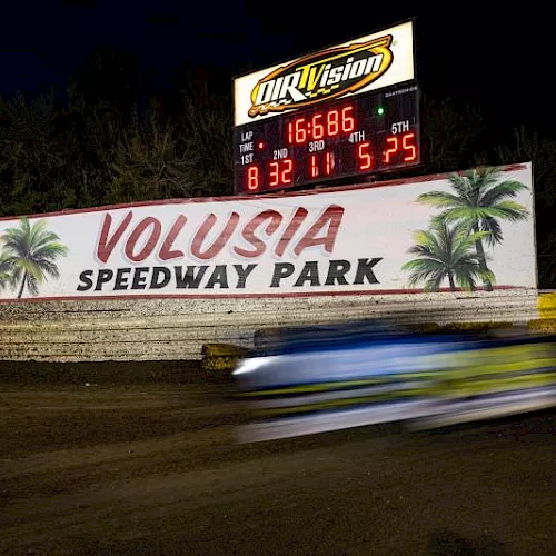 A night race at Volusia Speedway Park with a glowing scoreboard and a speeding car blurring past on the dirt track.