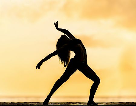 A person doing a dynamic yoga or dance pose at sunset, silhouette against a warm sky, stretching arms and legs dramatically.