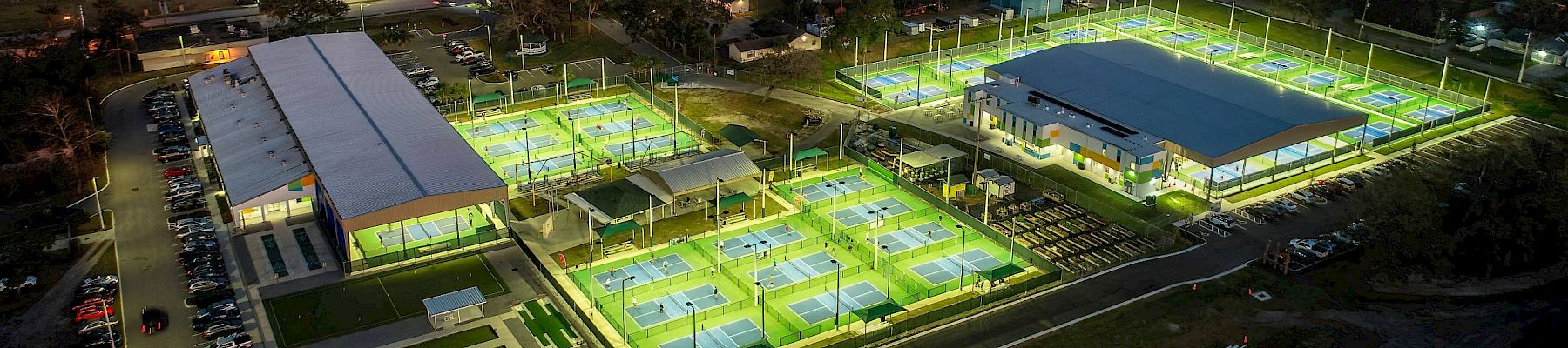 Aerial view of a sports complex with illuminated tennis courts and buildings at night, bordered by a road and water in the distance.