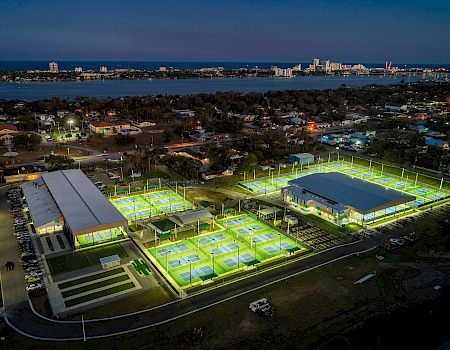 Aerial view of a sports complex with illuminated tennis courts and buildings at night, bordered by a road and water in the distance.