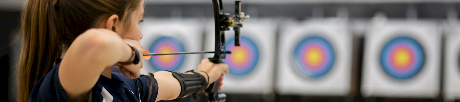A female archer aims a bow at indoor targets, arrows nocked, focusing on targets with concentric rings in the background.