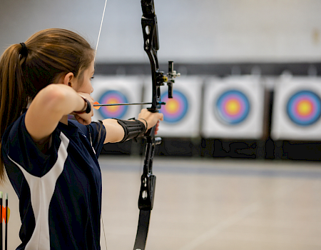 A female archer aims a bow at indoor targets, arrows nocked, focusing on targets with concentric rings in the background.