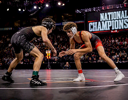 Two wrestlers face off on a mat during a National Championship match, with a crowd watching as the sign reads &ldquo;NATIONAL CHAMPIONSHIP.&rdquo;