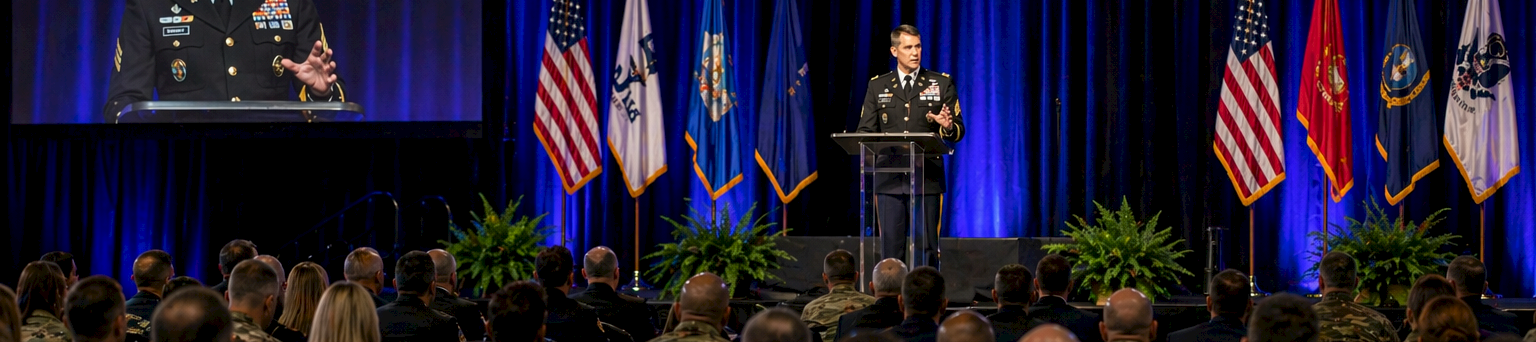 A uniformed speaker addresses a large audience in a dimly lit conference hall, with U.S. flags and blue curtains in the background, plus a screen.