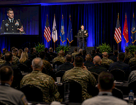 A uniformed speaker addresses a large audience in a dimly lit conference hall, with U.S. flags and blue curtains in the background, plus a screen.