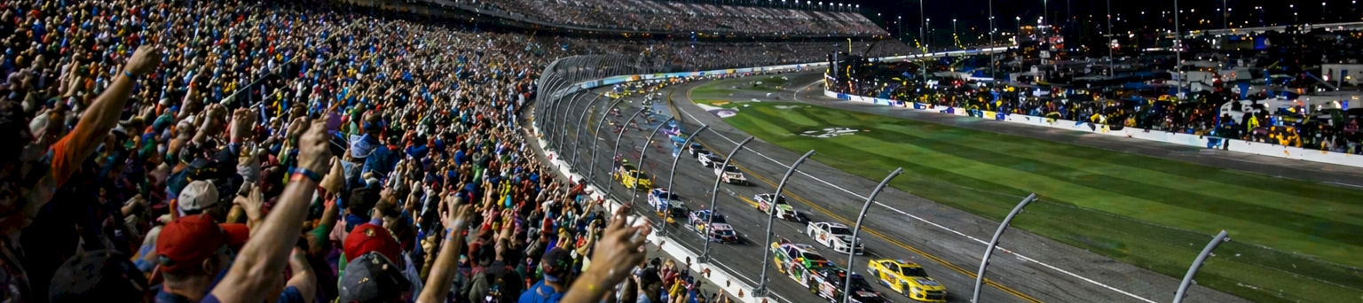 A packed race track at night with a roaring crowd, multiple cars on the oval, and cheering fans along the fence under bright lights.