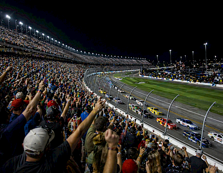 A packed race track at night with a roaring crowd, multiple cars on the oval, and cheering fans along the fence under bright lights.