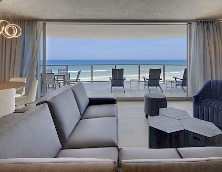 A modern living room with a beach view: grey sectional sofa, armchairs, coffee tables, and an open wall to a balcony overlooking the ocean.