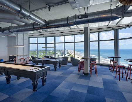 A bright, modern lounge with large windows, blue checkered carpet, pool table, high ceilings, and red stools around high tables, overlooking the ocean.