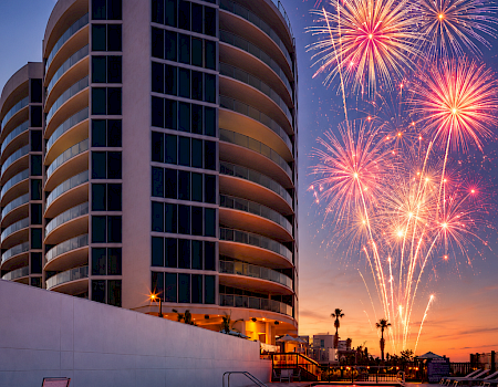 Fireworks burst over a modern high-rise hotel and its glowing pool at dusk, reflecting in the water.