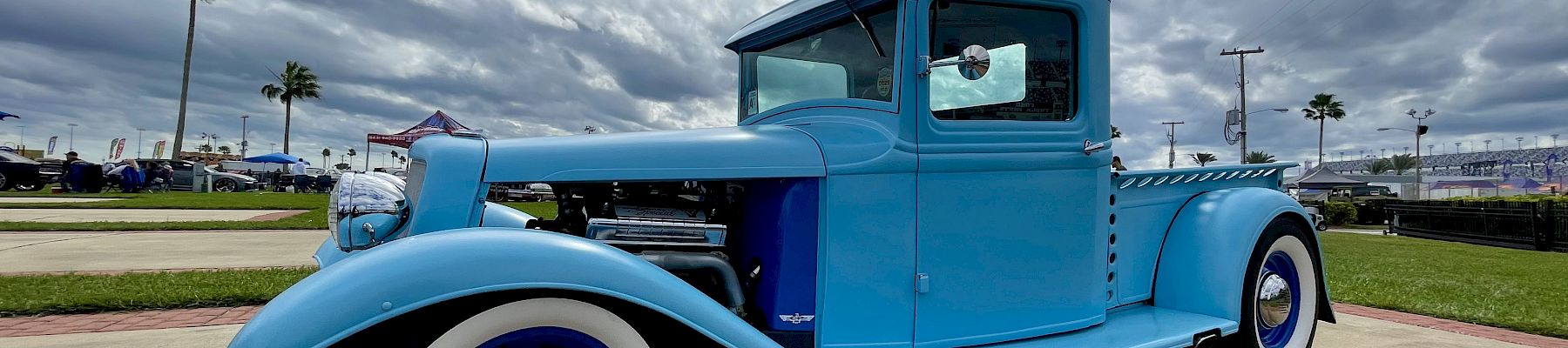 A bright blue vintage truck at a car show under cloudy skies, with palm trees and booths in the background.