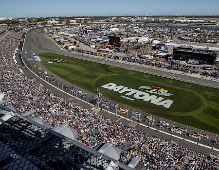 An aerial view of Daytona International Speedway during a packed NASCAR race, with cars on the track and fans filling the stands.