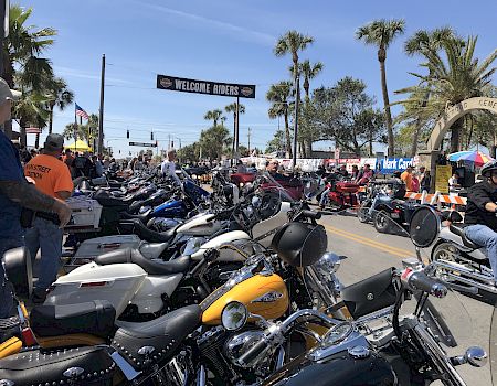 A large group of motorcyclists gathered on a sunny street under a “Welcome Riders” banner, with palm trees lining the road.