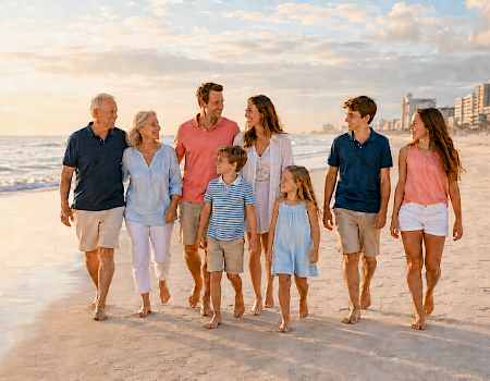 A family of six walks barefoot on a sunny beach at sunset, smiling and enjoying the seaside together.