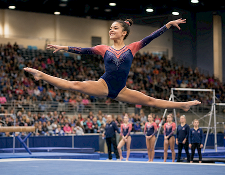 A gymnast in a blue leotard performs a balance pose in a packed indoor arena, arms outstretched.