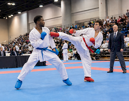 Two karate athletes in white gi with red/blue gloves spar in a crowded gymnasium arena, facing off during a match.