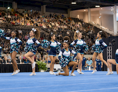 Cheer squad performs on an arena floor with blue-and-white pom-poms while a crowd watches.