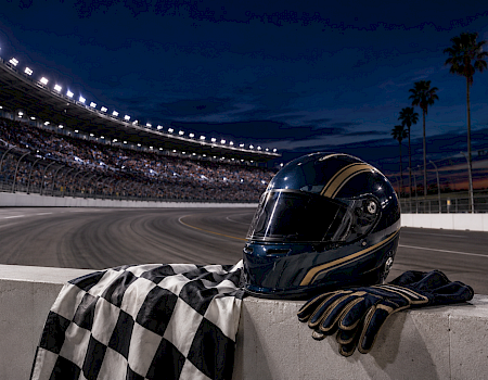 A driver’s helmet rests by a checkered flag on a night racetrack beside a roaring stadium.