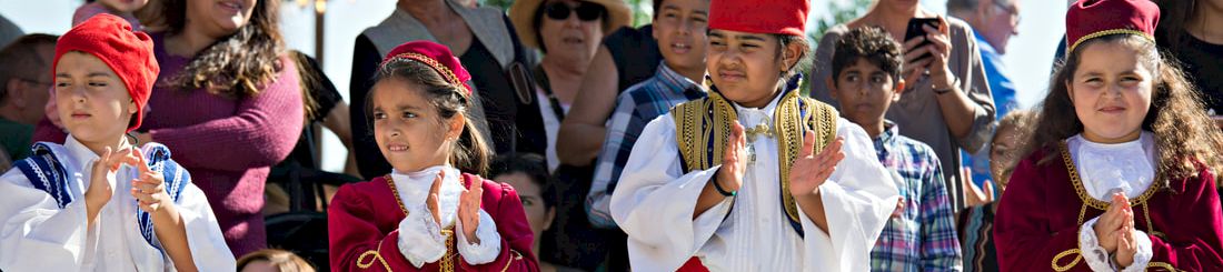 Children in folk costumes wearing red hats and blue dresses stand outdoors, clapping, as spectators watch in a sunny parade.
