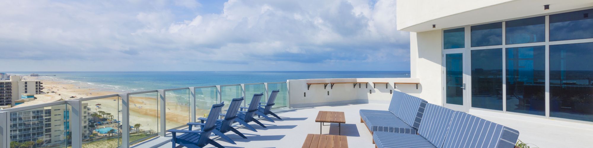 A sunny oceanfront balcony with blue-striped lounge chairs, a sofa, and a view of the sandy beach and clouds.