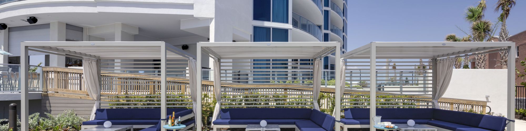 Modern hotel terrace with blue daybeds under a pergola, small tables, and a neatly kept lawn beside a glass-and-white building.