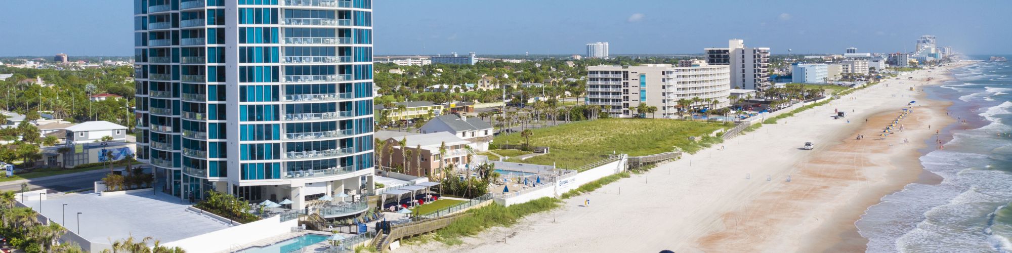 Aerial view of a beachfront resort with tall hotel buildings, palm trees, and rows of blue umbrellas along a sandy shore.