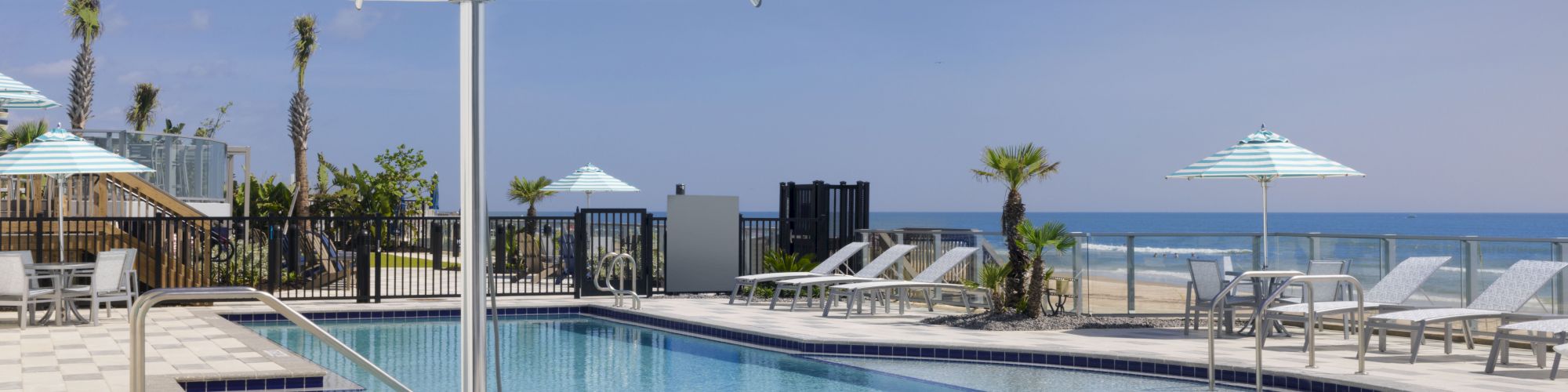 A beachfront resort pool with striped teal-and-white umbrellas, lounge chairs, and the ocean in view.