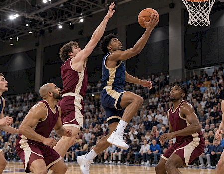 A basketball player leaps for a shot as defenders vie in a packed indoor arena, with the hoop overhead.