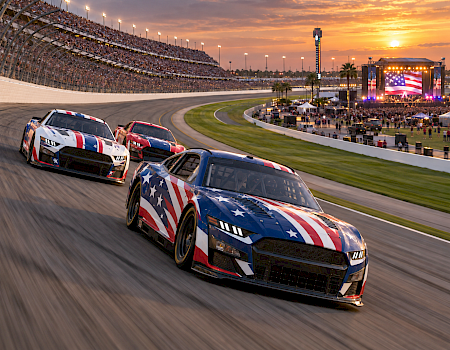 Three American-flag race cars speed around a stadium track at sunset, with cheering stands and a big stage in view.