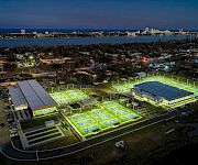 Aerial view of lit tennis courts by the coast at dusk, with a city skyline across the water.
