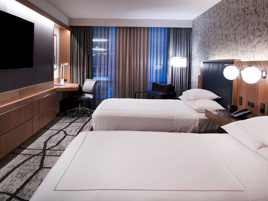 This image shows a modern hotel room with two neatly made beds, a large flat-screen TV, a desk with a chair, and contemporary lighting fixtures.