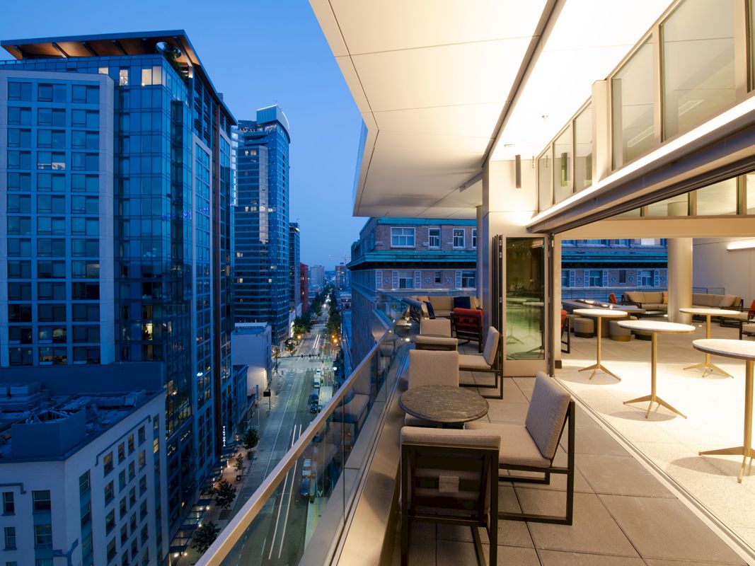 The image shows a modern balcony overlooking a cityscape with tall buildings, featuring seating and tables under evening lighting.