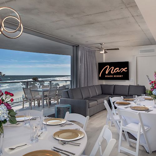 A beachside dining area with round tables, floral arrangements, set with gold-rimmed plates, a couch, and a view of the ocean, is depicted.