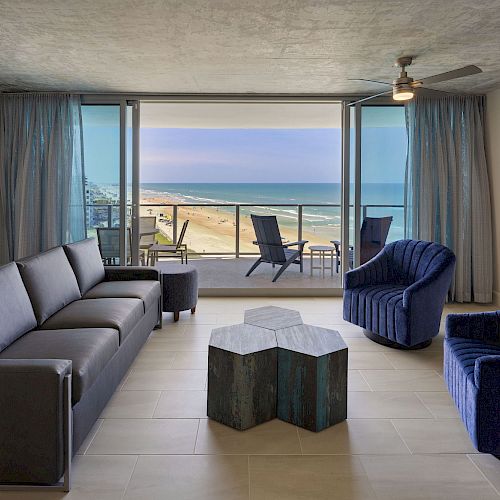 A modern living room with a beach view, featuring a black sofa, blue chairs, a unique wooden coffee table, and a TV on the wall.