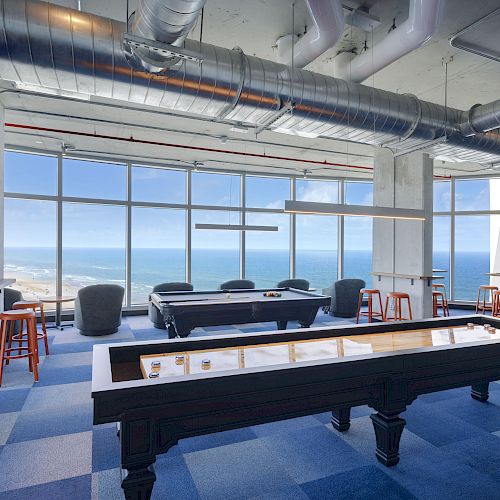 The image shows a modern game room with a shuffleboard table, pool table, seating areas, and large windows offering an ocean view.