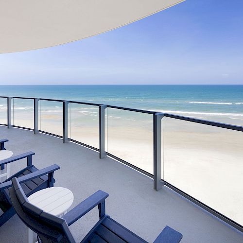 The image shows a balcony with blue chairs and small round tables overlooking a sandy beach and an expansive ocean under a clear sky.