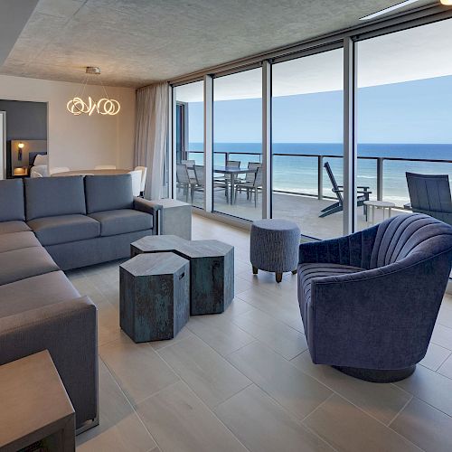 A modern living room with a large sectional sofa, blue armchair, unique coffee tables, floor-to-ceiling windows, and a view of the ocean.