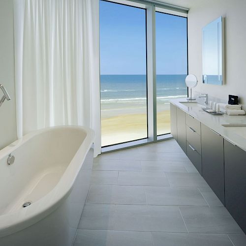 A modern bathroom features a bathtub, double sinks, large mirrors, and a window with an ocean view, complete with sleek, minimalist design elements.