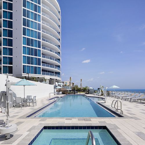 The image shows an outdoor pool area beside a modern high-rise building with glass windows, under a clear sky, with lounge chairs and umbrellas.