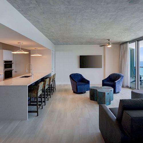 Modern open-plan living area with a kitchen on the left, seating area on the right, and large windows showcasing an ocean view.