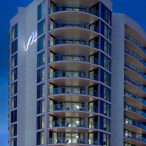 The image shows a modern high-rise building with multiple balconies and a sleek design, illuminated against a twilight sky, featuring a logo on the façade.