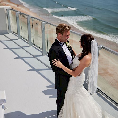 A couple in wedding attire stands on a balcony overlooking the ocean, embracing each other under the sun with a beautiful beach view.