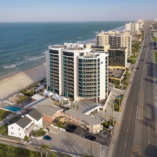 A coastal cityscape featuring a beachfront high-rise, adjacent buildings, and a road running parallel to the shoreline, extending into the distance.