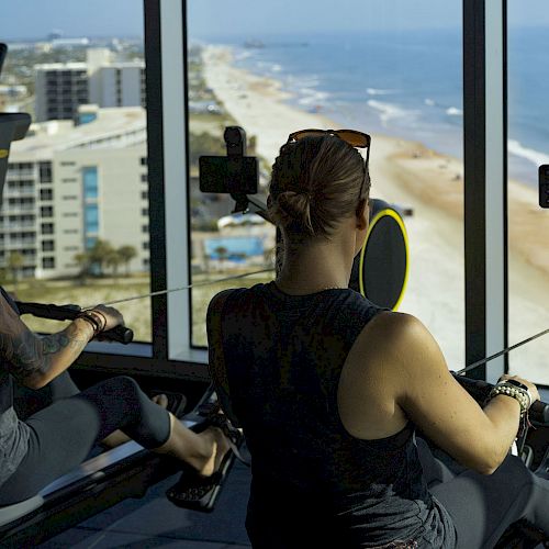 Two people are using rowing machines in a gym with large windows, offering a scenic beach view outside, enhancing their workout experience.